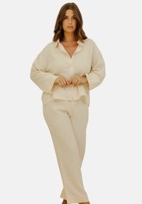 Femme aux cheveux bruns portant une chemise crème ample à manches longues avec boutons et un pantalon large assorti, debout devant un fond blanc.