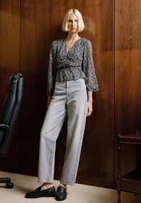 Crna bluzica s uzorkom i napuhanim rukavima, uparena s tamnosivim trapericama visokog struka ravnog kroja i crnim teksturiranim mokasinkama. Pozadina od drvenih panela.