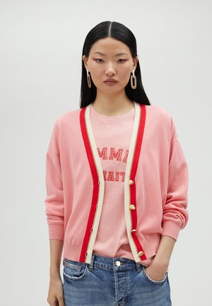 Pink cardigan with red trim and cream buttons, paired with a matching pink shirt featuring red text, denim jeans, and gold hoop earrings.