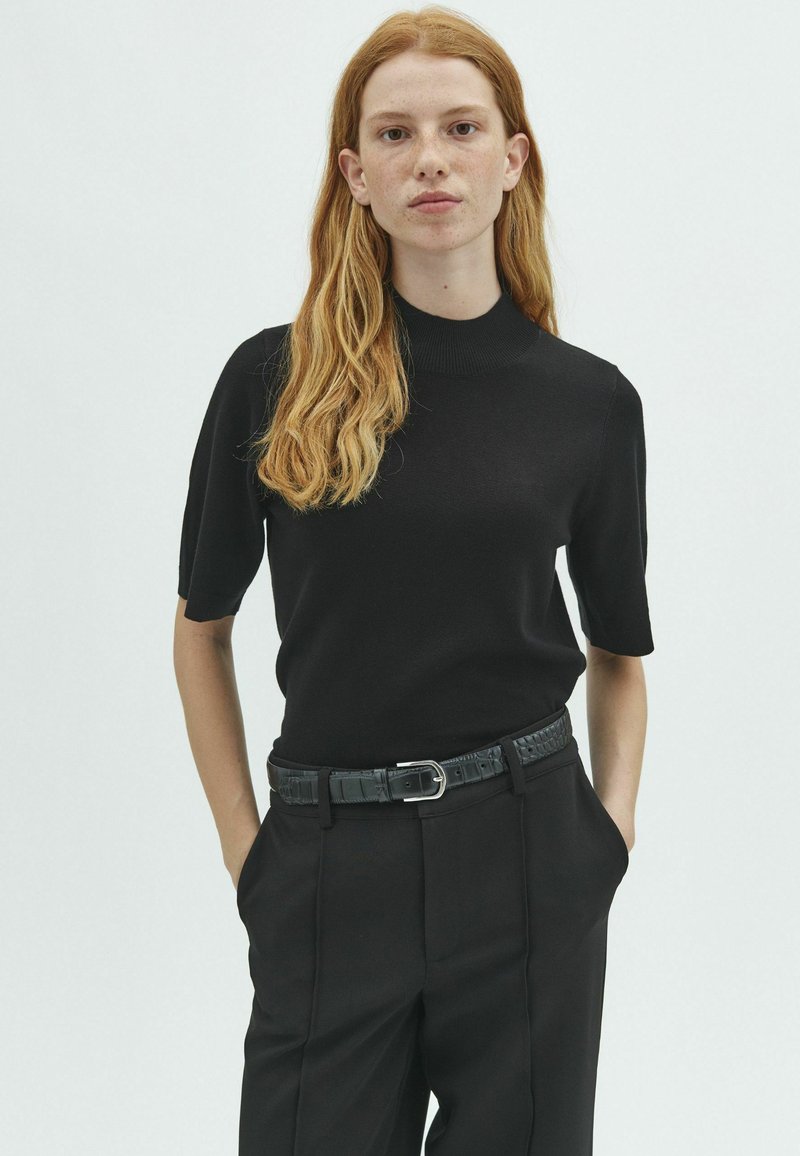 Jeune personne aux longs cheveux roux portant un haut noir moulant à manches courtes et un pantalon noir avec ceinture, debout les mains dans les poches.