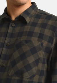 Chemise en flanelle à carreaux vert foncé et bleu marine. Présente un col, des boutons de fermeture et une poche poitrine avec des détails de couture visibles.