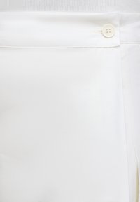 Gros plan sur la ceinture d'un pantalon blanc avec un petit bouton blanc et une couture visible sur un tissu lisse.