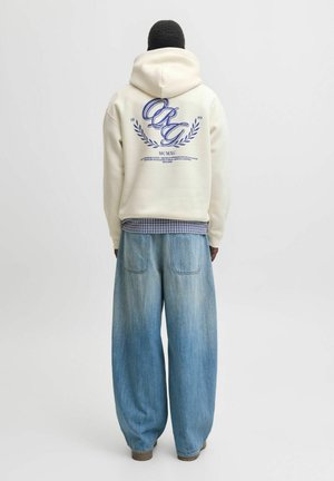 Personne portant un sweat à capuche crème avec un texte manuscrit bleu et un motif de laurier dans le dos, une chemise à carreaux en dessous et un jean ample bleu clair.