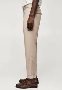 Beżowe spodnie damskie o dopasowanym kroju, z prostym przodem i mankietami u dołu. Noszone z brązowymi skórzanymi mokasynami o gładkiej fakturze i minimalistycznym designie.