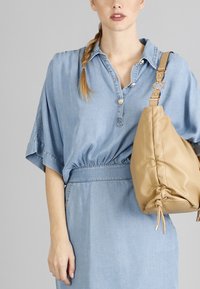 Camicetta di jeans azzurro chiaro con maniche corte, dotata di colletto abbottonato e vita elastica, abbinata a una borsa in morbida pelle color camoscio con cordoncino.