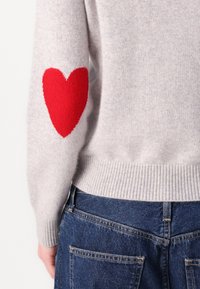 Maglione grigio a maglia con orlo e polsini a coste, caratterizzato da un grande cuore rosso sul gomito sinistro. Abbinato a jeans in denim blu.