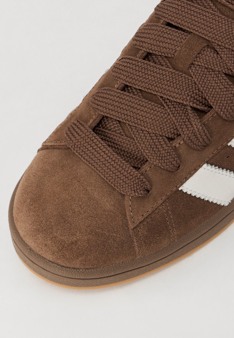 Zapatillas de ante marrón con puntera redondeada, cordones marrones texturizados y rayas blancas a lo largo del costado; suela de goma.