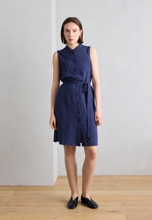 Γυναίκα που στέκεται μέσα σε κλειστό χώρο, φοράει αμάνικο μπλε σκούρο φόρεμα με λεπτές ρίγες, κουμπιά και ζώνη στη μέση, καθώς και μαύρα παπούτσια χωρίς κορδόνια.