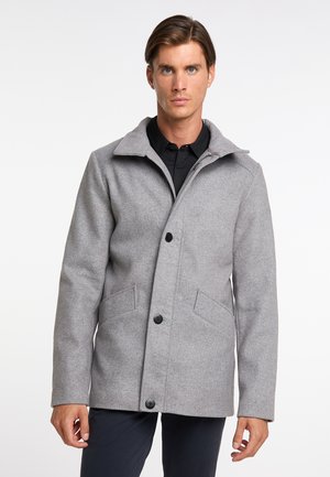 Uomo che indossa un cappotto grigio chiaro con bottoni e tasche sopra una camicia nera, in piedi davanti a uno sfondo bianco semplice.