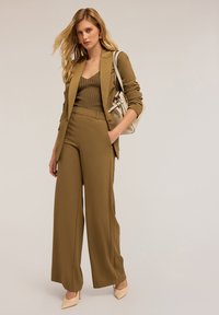 Khaki-kleurig op maat gemaakt pak met een gestructureerde blazer en een broek met wijde pijpen, gecombineerd met een geribbelde olijfgroene tanktop en naaktkleurige hakken.