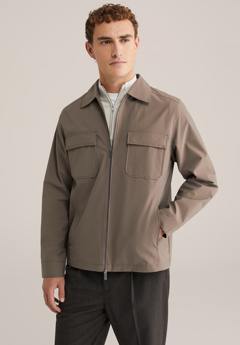 Homme aux cheveux bouclés courts portant une veste taupe zippée avec des poches poitrines à rabat et un pantalon gris foncé, regardant légèrement vers la droite.