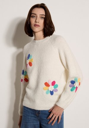 Žena s hnědými vlasy po ramena má na sobě bílý chlupatý svetr zdobený barevnými pletenými květinovými nášivkami a modré džíny.