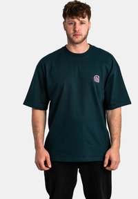 Camiseta de algodón verde oscuro con mangas cortas, cuello redondo y un pequeño logo rosa en el lado izquierdo del pecho. Ajuste casual, textura suave.