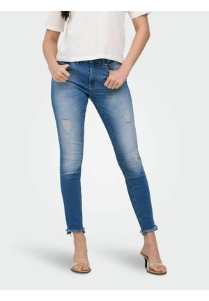 Jean slim bleu usé avec bas effilochés aux chevilles, porté avec des sandales à talons beiges et un haut blanc à manches courtes.