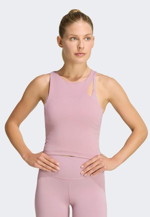 Kvinde med blondt hår i en hestehale iført en lyserød ærmeløs crop top og matchende leggings, stående med hænderne på hofterne.