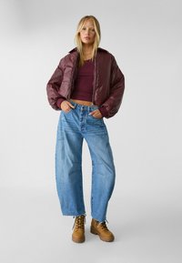 Bordeauxfarbene, kurze Pufferjacke mit langen Ärmeln, kombiniert mit hellblauen, weit geschnittenen Jeans mit Steppdetails und braunen Schnürboots.