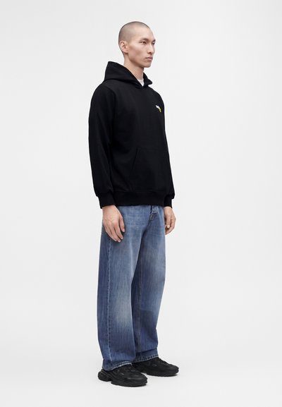Homme portant un sweat à capuche noir, un jean bleu délavé et ample, et des chaussures noires, debout devant un fond blanc uni.
