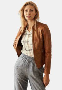 Bruine leren jas met een aansluitend ontwerp, voorzien van ritszakken en geribbelde accenten, gedragen over een licht geruit overhemd met knopen en grijze broek.