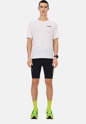 White short-sleeve athletic shirt, black compression shorts, bright yellow socks, and green running shoes with black accents. Minimal branding visible.