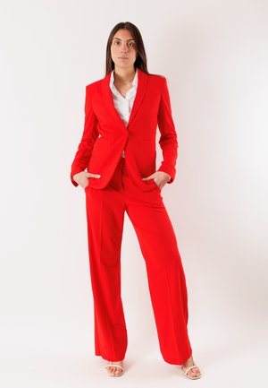 Donna in piedi con le mani nelle tasche, indossa un completo rosso vivo con pantaloni a gamba larga, una camicia bianca e sandali beige con punta aperta.