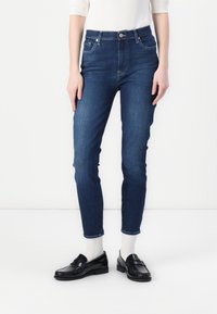 Jeans de mezclilla azul oscuro con un ajuste slim, cintura alta, diseño de cinco bolsillos y detalles de costura mínimos. Combinados con mocasines negros y calcetines blancos.