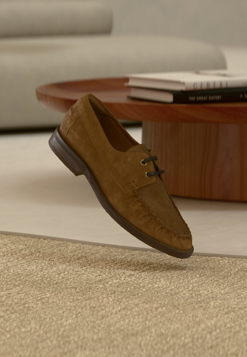 Mocassin en daim marron avec des lacets noirs flottant au-dessus d'un tapis beige texturé, avec une table ronde en bois et des livres en arrière-plan.