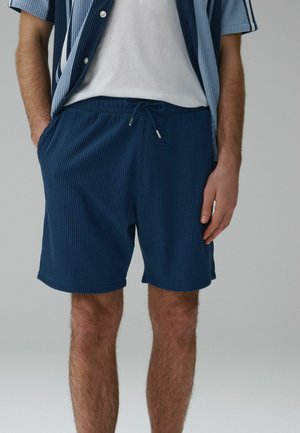 Mann trägt marineblaue, gerippte Shorts mit Kordelzug und ein passendes kurzärmeliges Hemd über einem weißen T-Shirt, steht mit entspannten Händen.