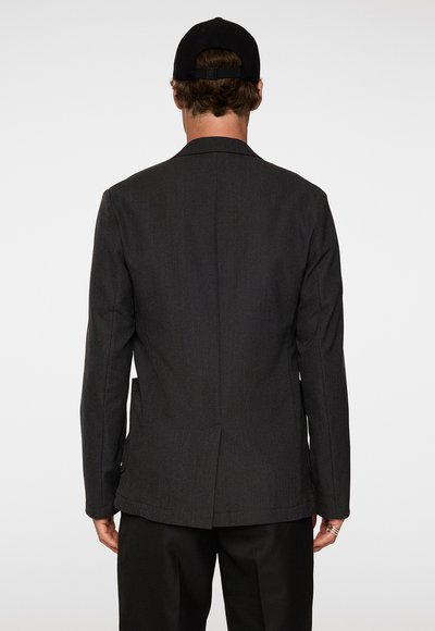 Blazer grigio scuro con una vestibilità su misura, revers a punta, una fessura singola sul retro e due tasche laterali. Indossato sopra pantaloni neri.