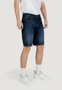 Shorts en denim bleu foncé avec des coutures orange, poches avant et une taille standard. Portés avec un t-shirt blanc et des baskets.