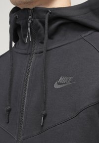 Närbild av en person som bär en svart Nike zip-up hoodie med snören och en hög krage som täcker nacken.