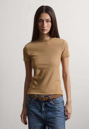 Frau mit langen dunklen Haaren, die ein beiges, tailliertes Kurzarmshirt trägt, das in eine blaue Jeans mit braunem Gürtel gesteckt ist, stehend vor einem schlichten Hintergrund.