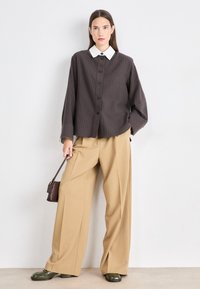 Brown textured button-up shirt with a white collar, paired with wide beige trousers and dark green shoes. Small brown shoulder bag detail.