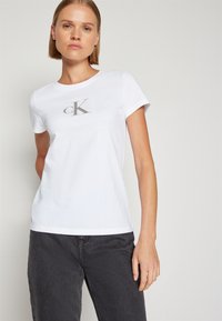 Biela bavlnená tričko s krátkymi rukávmi a okrúhlym výstrihom. Na prednej strane je decentne vytlačené šedé logo cK. Hladká textúra.