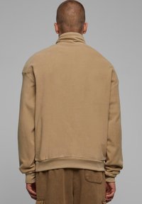 Beige fleecegenser med høy krage, ribbestrikkede mansjetter og hem. Glatt tekstur med oversized passform, vist fra baksiden.