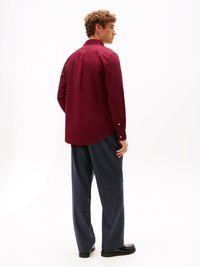 Bordeaux shirt met een kraag en lange mouwen; geplooid detail op de achterkant. Gepaard met een losse, donkere broek en zwarte schoenen.