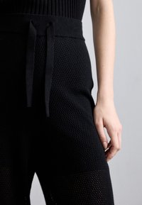 Pantaloni jogger neri in mesh con superficie testurizzata, dotati di una cintura ampia e chiusura con lacci. Mani posizionate vicino alla vita.