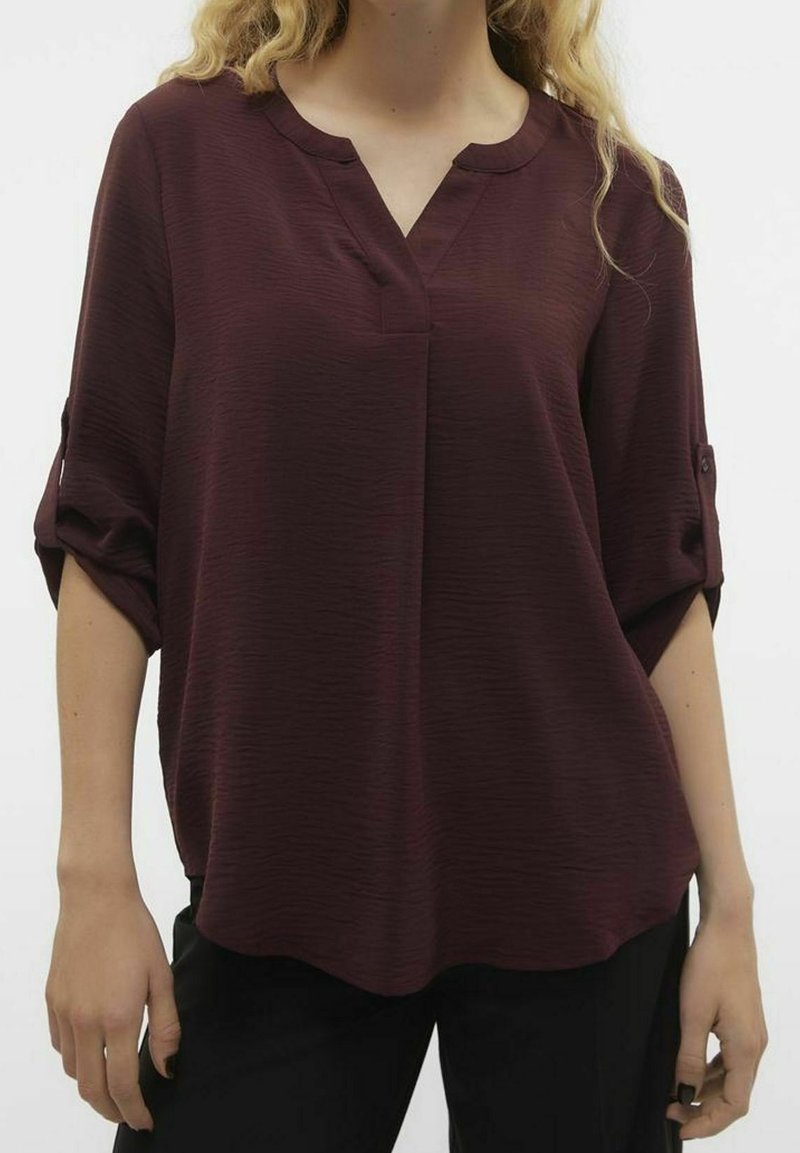 Blusa bordeaux con scollo a V e maniche arrotolate, realizzata in tessuto testurizzato; vestibilità comoda con orlo leggermente più lungo sul retro.