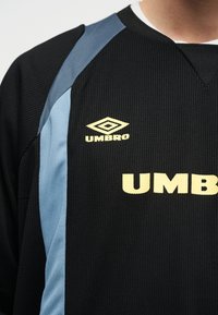 Črna športna majica z modrimi stranskimi paneli in rumenim logotipom Umbro ter napisom na prsih, nošena na zgornjem delu telesa osebe.