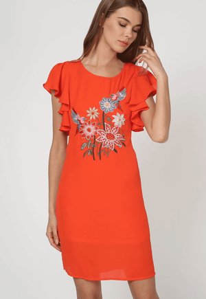 Mujer con un vestido naranja brillante con mangas cortas fruncidas y bordados florales con colibríes en el frente.