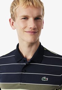 Polo blu navy con strisce orizzontali verdi e bianche, dotato di colletto, due bottoni e logo di coccodrillo ricamato verde sul petto.