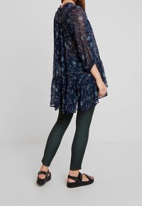 Námořnická modrá průsvitná šaty s květinovými vzory, volánkovým lemem a třičtvrtinovými rukávy, spárované s tmavě zelenými legínami a černými sandály.