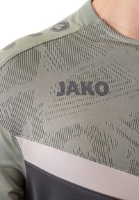 Grünes Sportshirt aus strukturiertem Stoff, mit einem dunkelgrauen Akzent und hellgrauem Streifen. Das "JAKO"-Logo ist gut sichtbar angebracht.