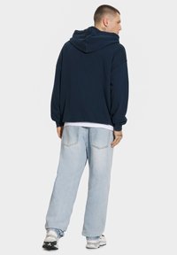 Granatowa bluza z kapturem o luźnym kroju, w zestawie z jasnoniebieskimi dżinsami. Materiał wydaje się miękki i gładki w dotyku.