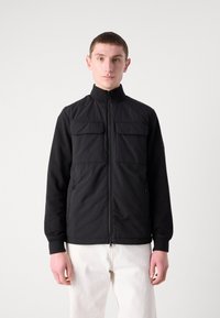 Jachetă neagră cu guler înalt, având două buzunare pe piept și buzunare laterale cu fermoar, fabricată dintr-un material texturat, asortată cu pantaloni albi.