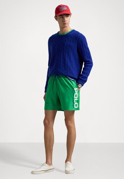Pull en tricot bleu, short vert avec logo "POLO", casquette rouge avec texte "Polo", baskets blanches, le tout présenté sur un fond neutre.