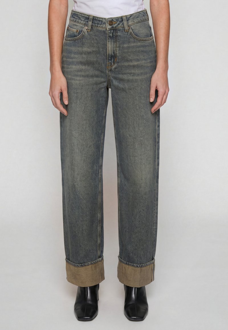 Personne portant un jean gris délavé à coupe droite avec de larges revers montrant un tissu intérieur beige et des bottes noires à bout carré.