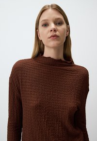 Pull en maille texturée marron avec un col montant et un bord effiloché subtil. Les manches longues et la coupe décontractée renforcent son apparence décontractée.