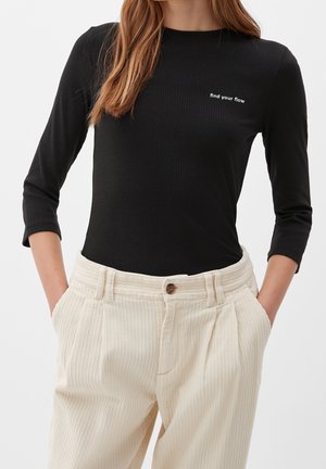Femme portant un t-shirt côtelé noir à manches longues avec le texte « find your flow » et un pantalon beige en velours côtelé taille haute, les mains dans les poches.