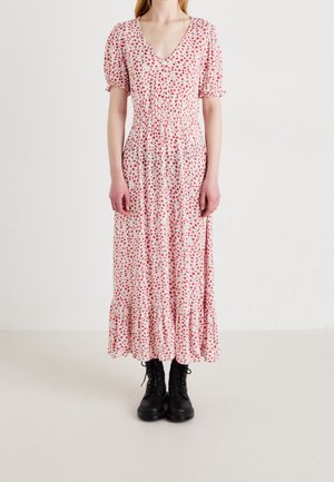 Mujer vestida con un vestido blanco midi con estampado floral rojo, mangas abullonadas, escote en V, bajo con volantes y botas negras con cordones hasta el tobillo, de pie contra una pared blanca.