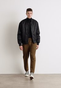 Schwarze Bomberjacke über einem schwarzen Rollkragenpullover, braune Hose und schwarz-weiße Sneakers. Glatte Texturen mit einem lässigen Design.